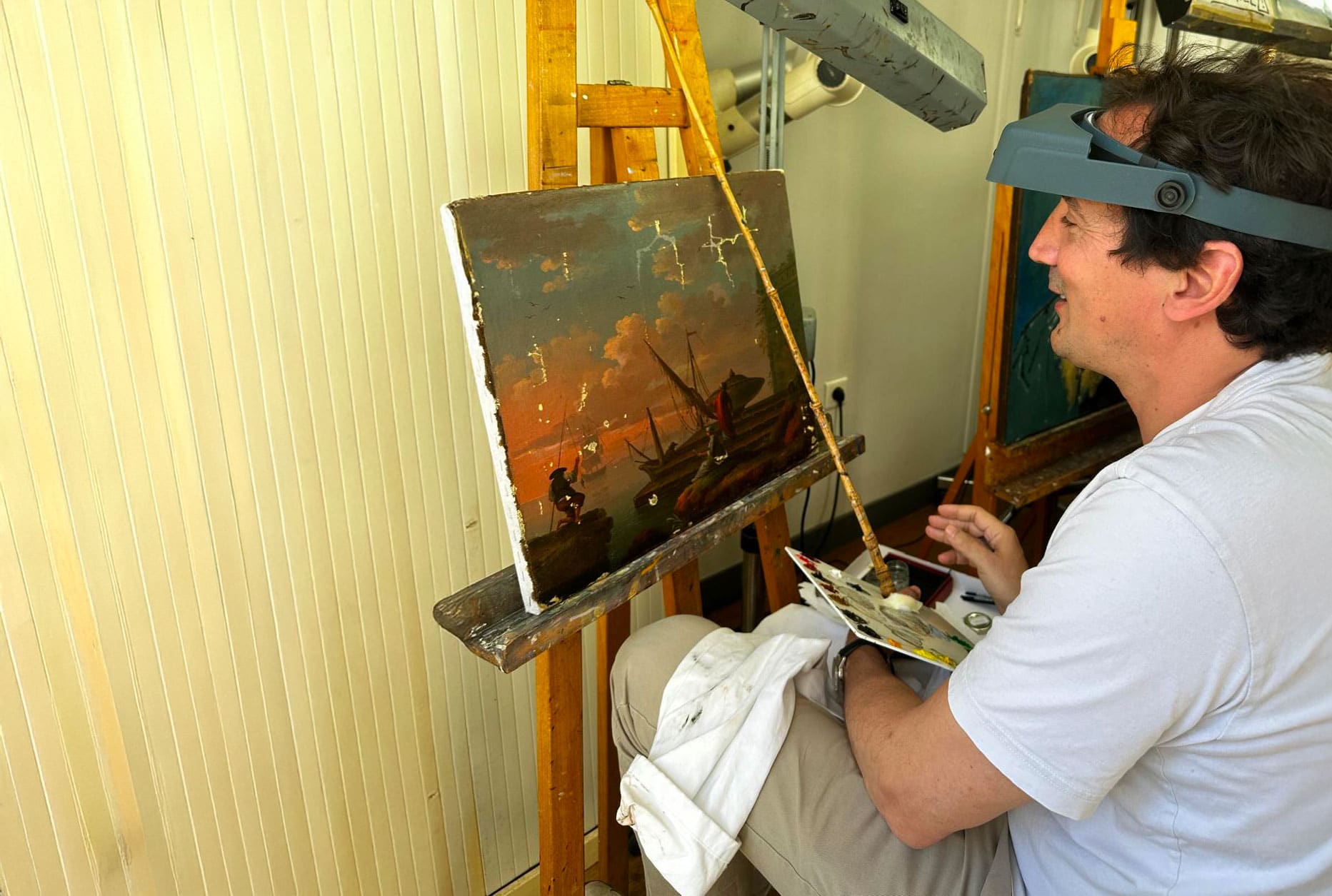 Matthieu Darrasse à l'atelier, loupe frontale et palette en main, devant une marine ancienne en cours de restauration